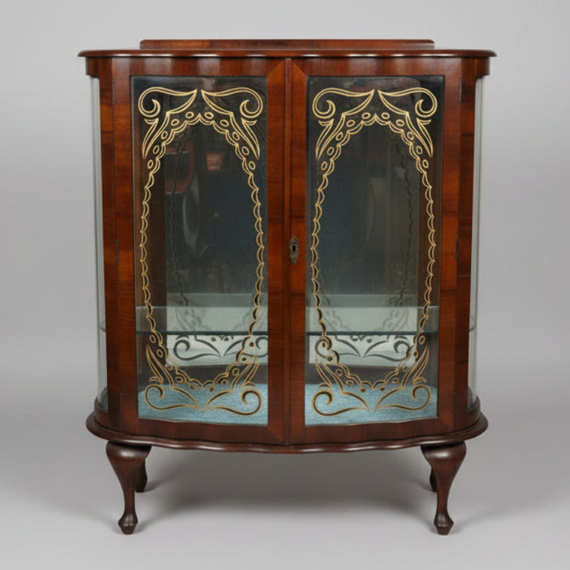 A curved wooden display cabinet with glass doors and ornate gold-toned metal scrollwork, photographed in what appears to be a furniture store or antique shop.