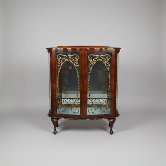 A curved wooden display cabinet with glass doors and ornate gold-toned metal scrollwork, photographed in what appears to be a furniture store or antique shop.