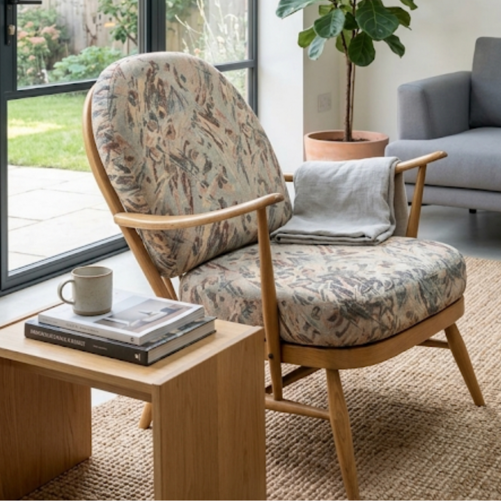 A mid-century modern armchair with a floral patterned cushion and light wood frame, styled in a bright living room next to a wooden side table and a potted fiddle-leaf fig plant.