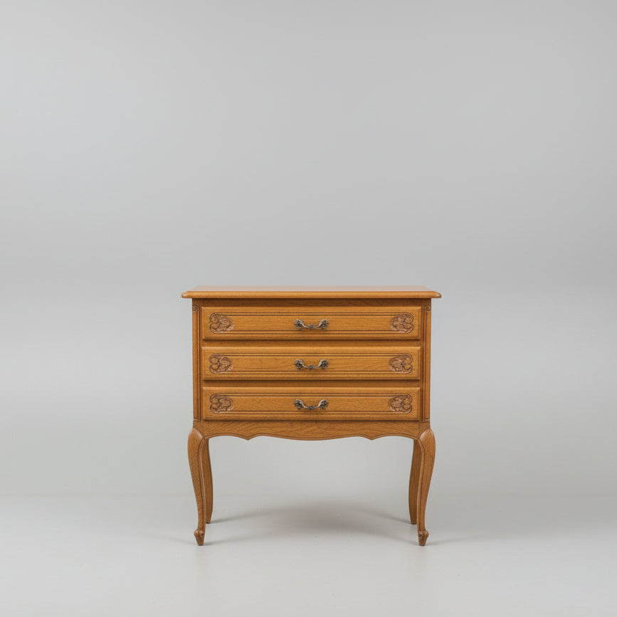 A three-drawer wooden chest of drawers with carved floral details on the drawer fronts and curved cabriole-style legs, photographed outdoors against a dark wooden wall and gravel ground.