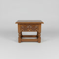 A dark brown wooden end table with carved floral details on the front panel and turned legs, photographed in what appears to be a warehouse or furniture store.