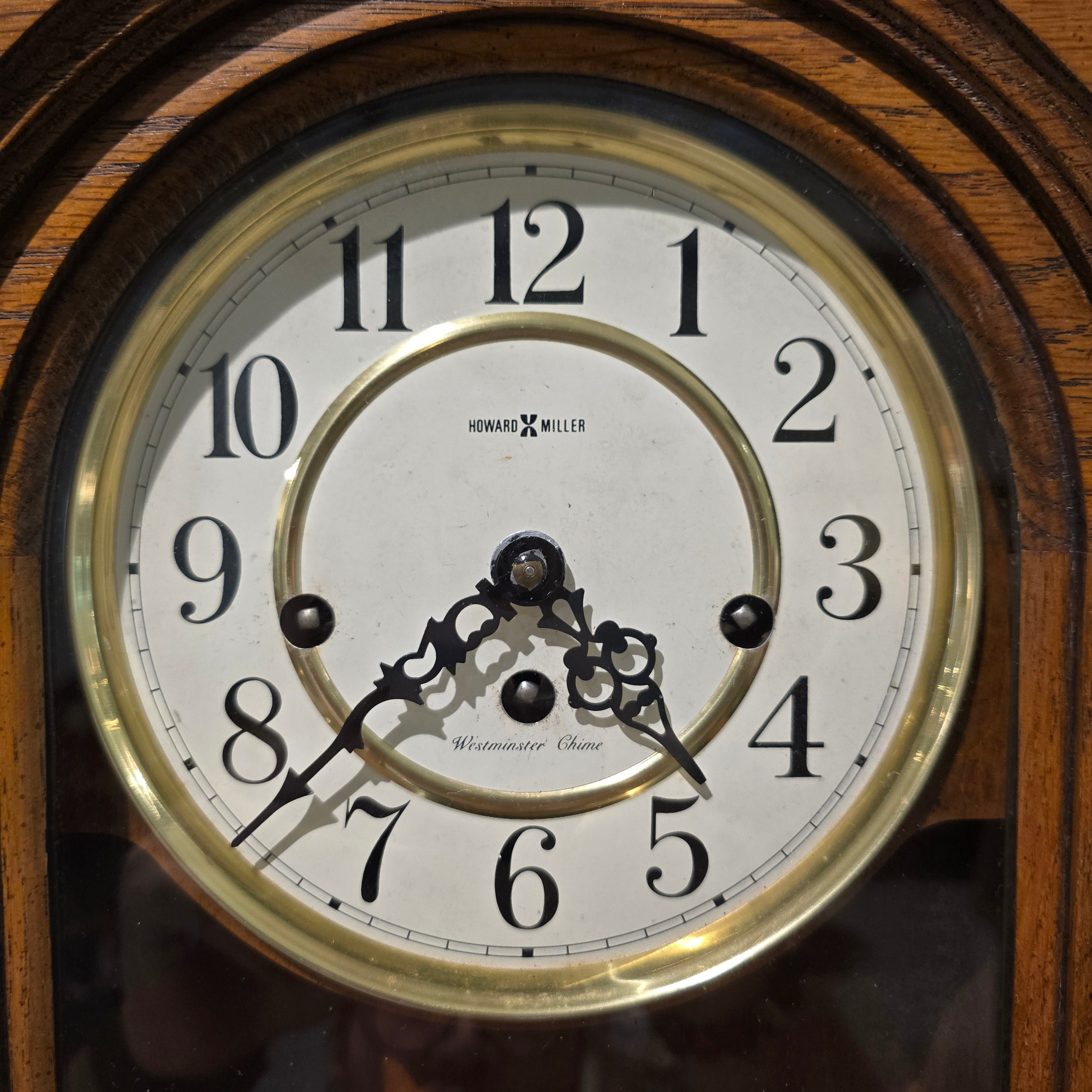 Close-up of a vintage wall clock face with a white dial, black Arabic numerals, ornate black hands, and a gold-toned inner ring, set within a dark wood case.