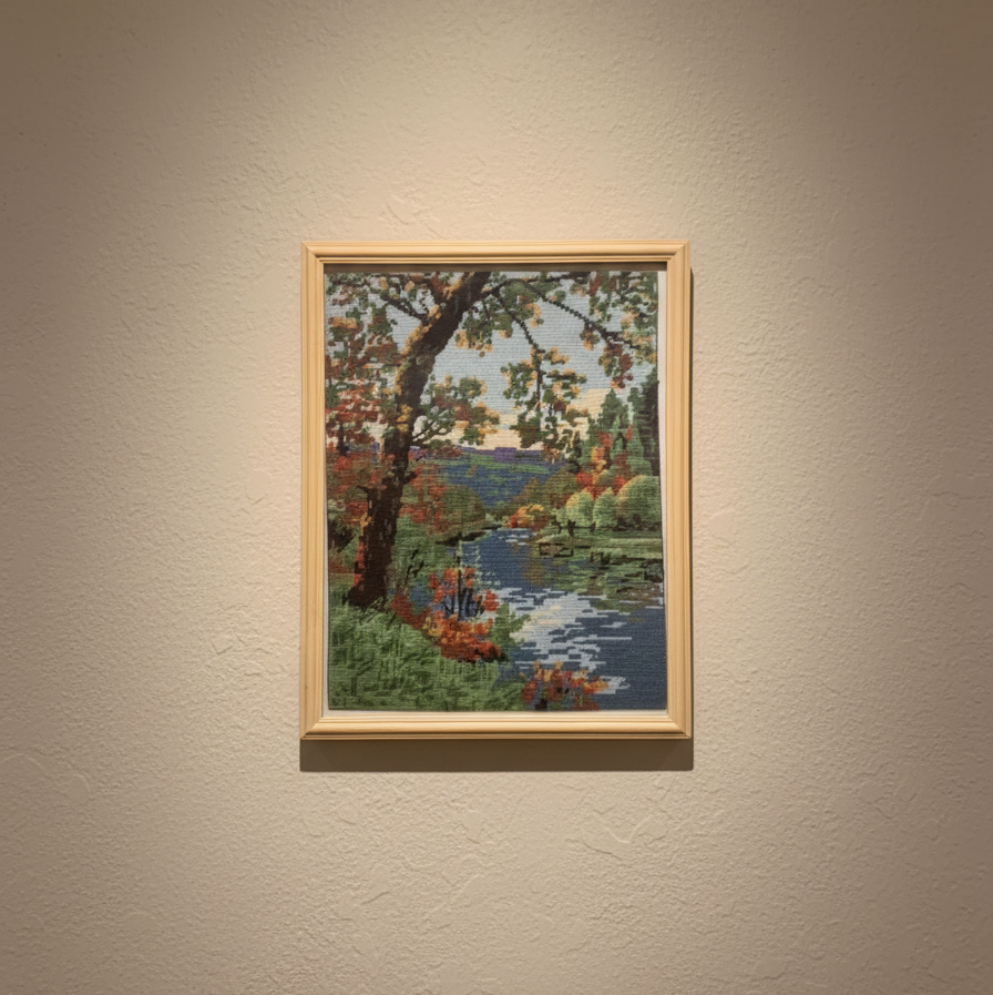 Framed needlepoint of a landscape with trees and water on a beige wall.