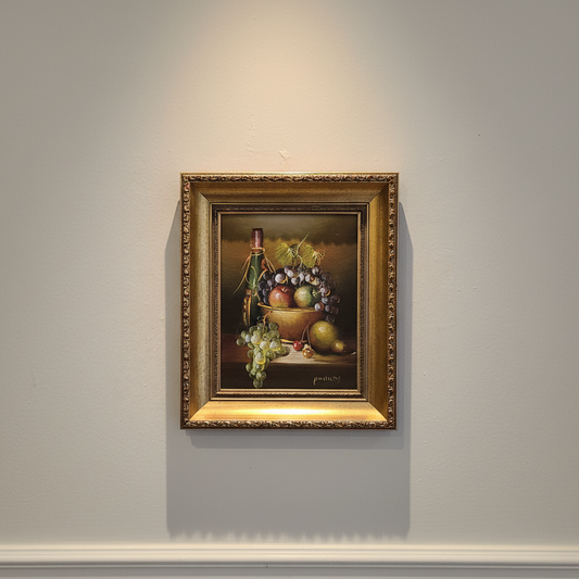 Framed still life painting of fruits and a bottle on a gray wall.