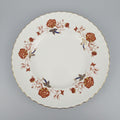 A white ceramic plate with a scalloped rim, decorated with a floral border pattern in terracotta, gold, and blue, shown on a dark wooden table.