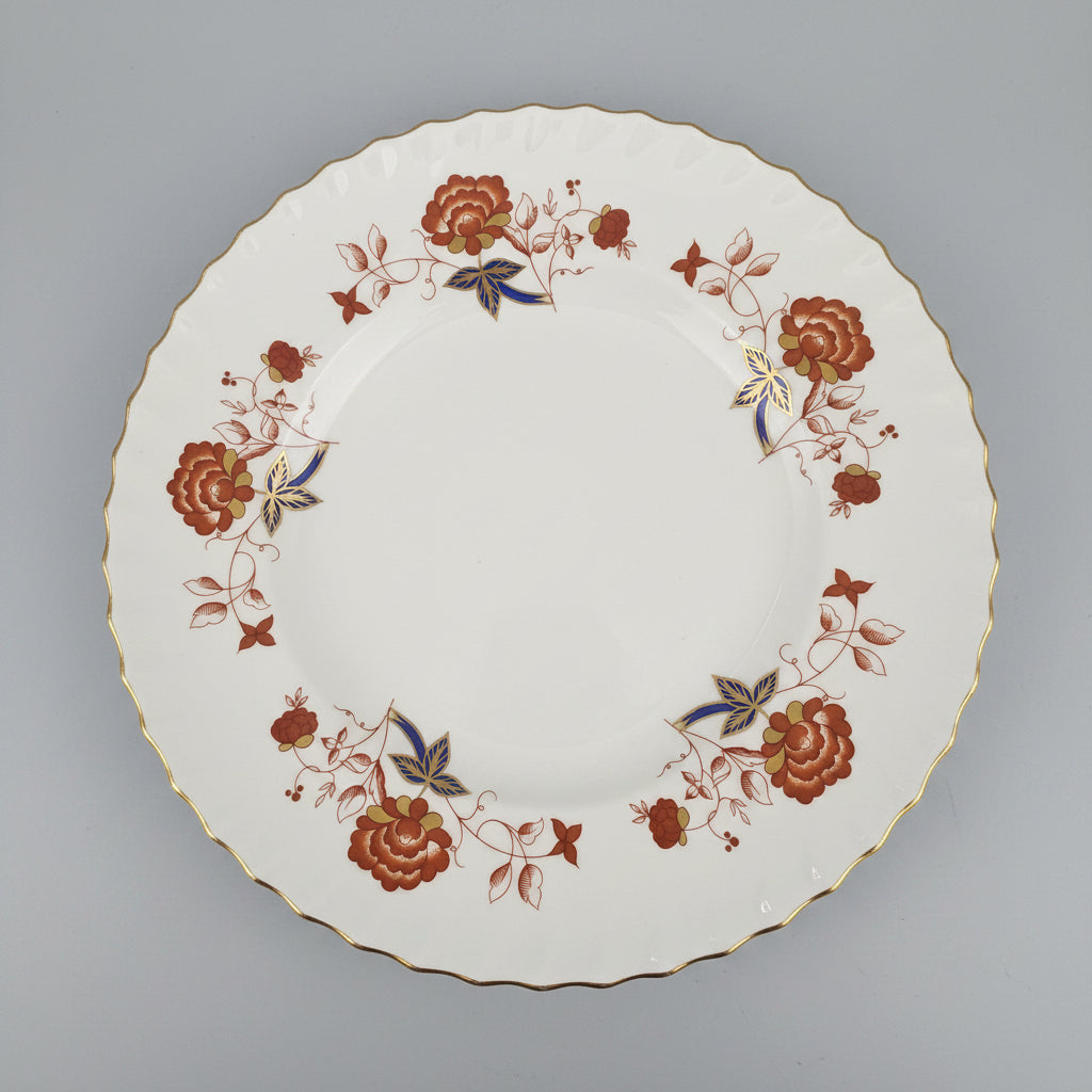 A white ceramic plate with a scalloped rim, decorated with a floral border pattern in terracotta, gold, and blue, shown on a dark wooden table.
