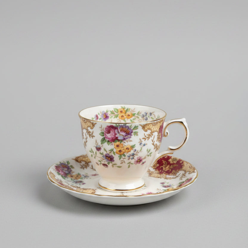 An ornate vintage-style floral teacup and saucer set with gold trim, displayed on a polished wooden table in a softly blurred antique shop interior.