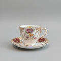 A vintage  teacup and saucer set with a white porcelain base, decorated with a colorful floral pattern and gold trim, resting on a dark brown textured surface against a plain beige wall.