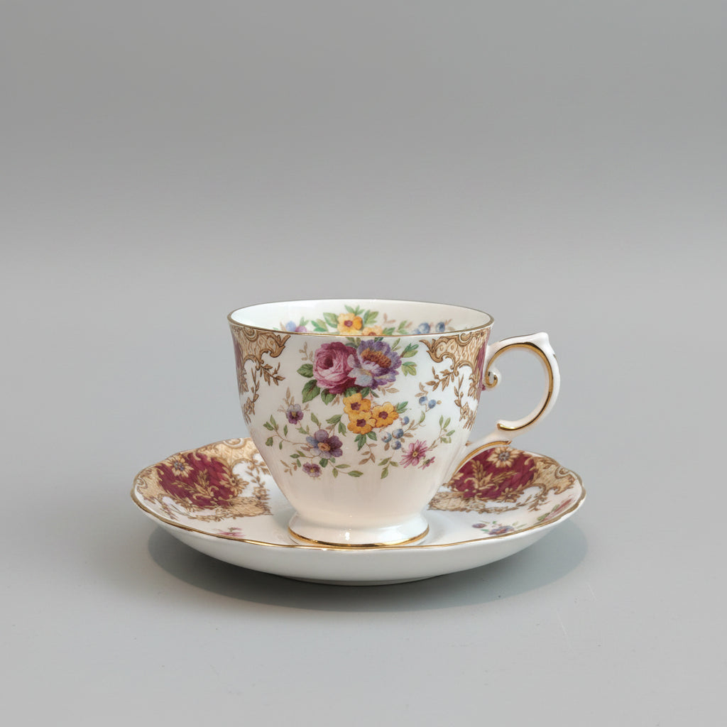 A vintage  teacup and saucer set with a white porcelain base, decorated with a colorful floral pattern and gold trim, resting on a dark brown textured surface against a plain beige wall.