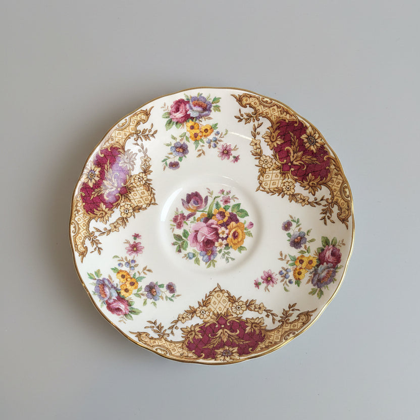 An ornate white porcelain plate with a floral pattern and gold trim, shown on a brown textured surface with decorative gold borders. A second matching plate is partially visible on the right.