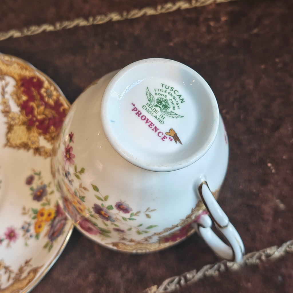 A close-up of a vintage porcelain teacup and saucer, showing the inside bottom of the cup with 'TUSCAN PROVENCE' printed in green and red. The white porcelain features delicate pink, red, and yellow floral patterns, resting on a dark brown textured surface with gold trim.
