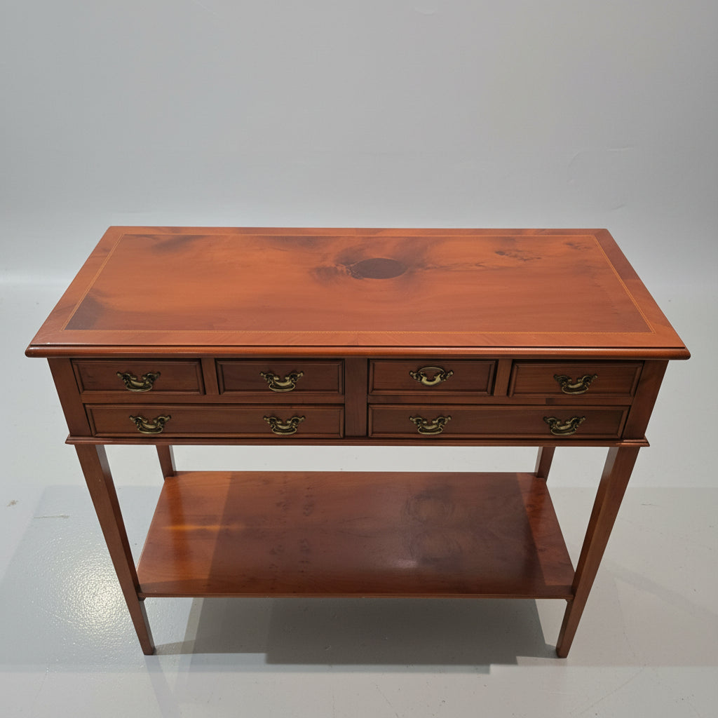 A reddish-brown wooden console table with six drawers, brass handles, and an open lower shelf, photographed in a furniture store with other items visible in the background.