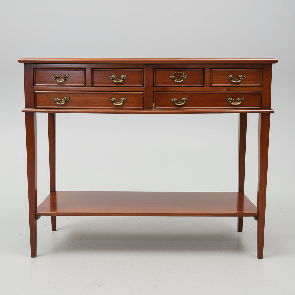 A dark reddish-brown wooden console table with six drawers, brass handles, and an open lower shelf, photographed in a furniture showroom.
