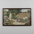 A framed needlepoint artwork depicting a vintage English countryside scene with a thatched cottage and a person on a bicycle.