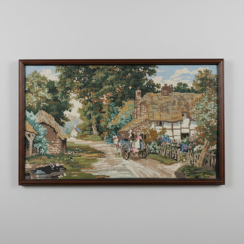 A framed needlepoint artwork depicting a vintage English countryside scene with a thatched cottage and a person on a bicycle.