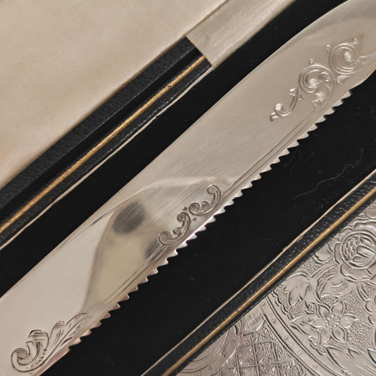 A close-up of a serrated bread knife with ornate floral engravings on the blade, resting in a black presentation box with a cream fabric cover.