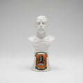 A white ceramic bust sculpture of a bald man with a mustache, displayed on a dark wooden surface against a textured off-white wall with a decorative gold-toned trim.