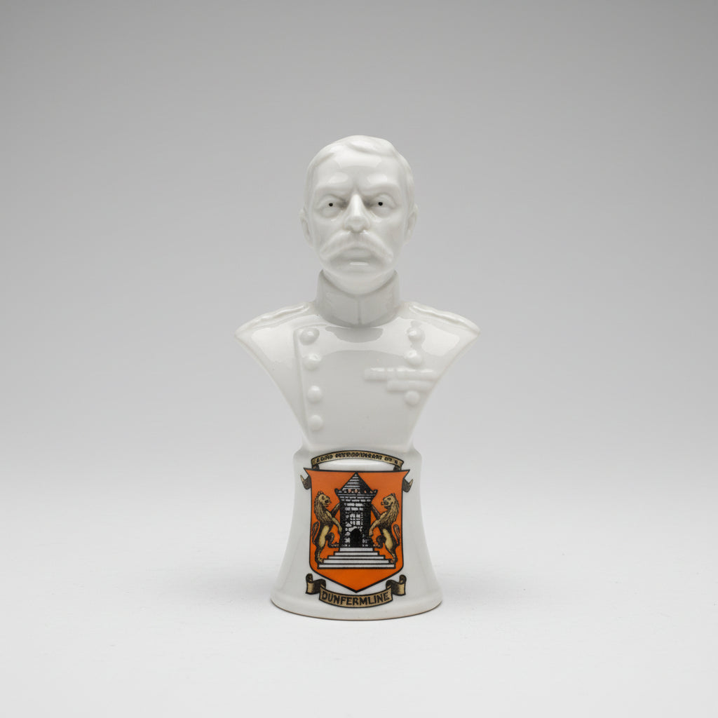 A white ceramic bust sculpture of a bald man with a mustache, displayed on a dark wooden surface against a textured off-white wall with a decorative gold-toned trim.