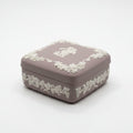 A hand holding a square Wedgwood-style ceramic box in a muted taupe-brown color with a raised cream cherub and grapevine border on the lid.