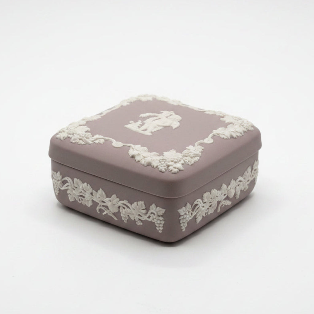 A hand holding a square Wedgwood-style ceramic box in a muted taupe-brown color with a raised cream cherub and grapevine border on the lid.