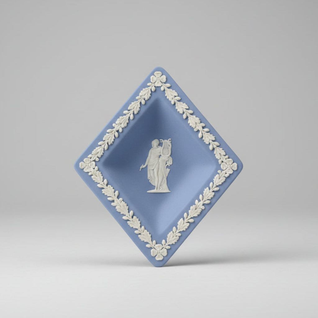 a diamond-shaped blue and white Wedgwood-style ceramic dish with a decorative border and a classical figure relief in the center, set against a blurred antique shop background.