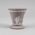 a classic Wedgwood-style ceramic cup in a dusty mauve-pink color with white relief decorations of a cherub and a laurel wreath, set against a blurred background of a furniture store.