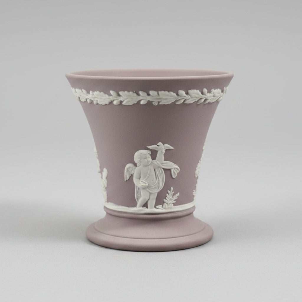 a classic Wedgwood-style ceramic cup in a dusty mauve-pink color with white relief decorations of a cherub and a laurel wreath, set against a blurred background of a furniture store.