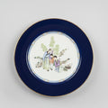 A round ceramic plate with a wide dark blue rim and a hand-painted central scene of figures in traditional clothing, displayed on a wooden table in an antique shop setting.