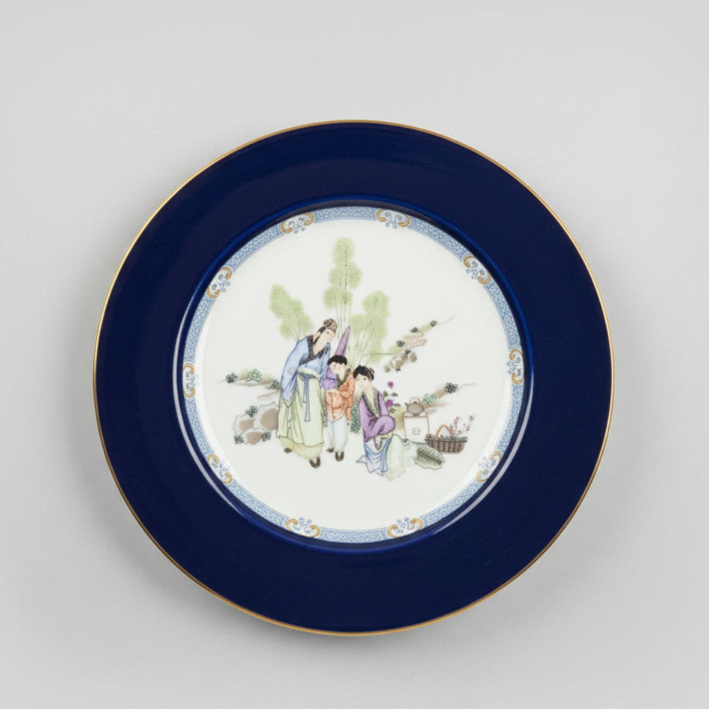 A round ceramic plate with a wide dark blue rim and a hand-painted central scene of figures in traditional clothing, displayed on a wooden table in an antique shop setting.
