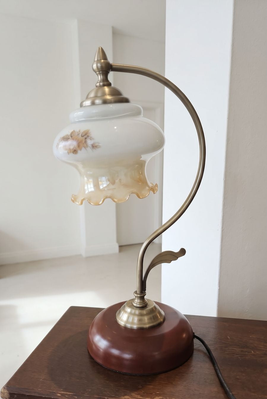 A vintage-style table lamp with a white glass shade decorated with gold floral motifs, mounted on a curved brass-colored metal arm with a dark reddish-brown wooden base, shown sitting on a dark wooden table.