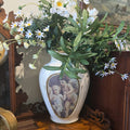 A white ceramic vase with a central classical cherub illustration, filled with white daisies, small purple wildflowers, and green leaves, displayed on a carved wooden console table.