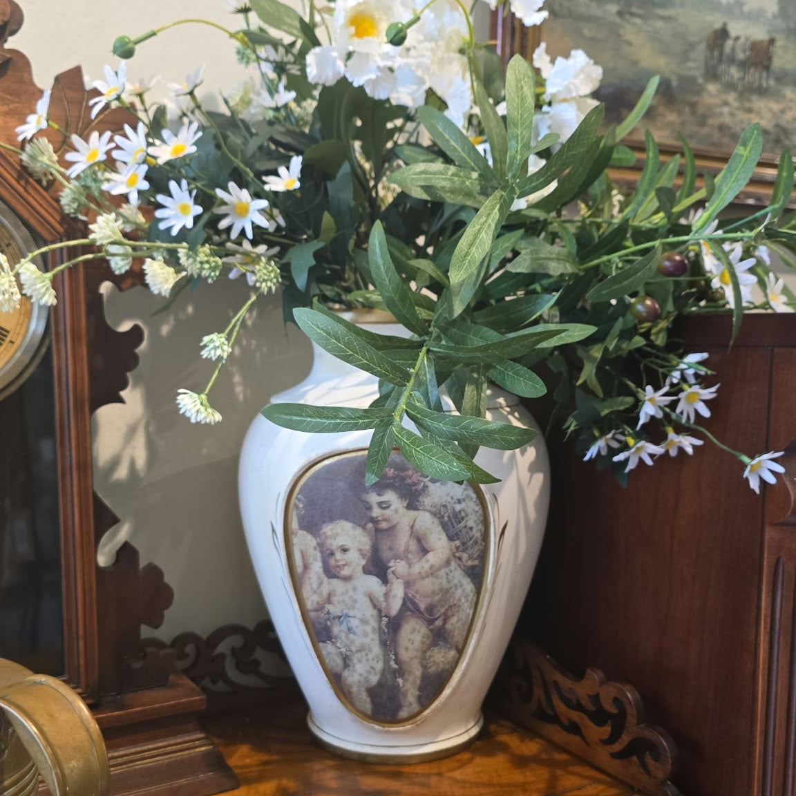 A white ceramic vase with a central classical cherub illustration, filled with white daisies, small purple wildflowers, and green leaves, displayed on a carved wooden console table.