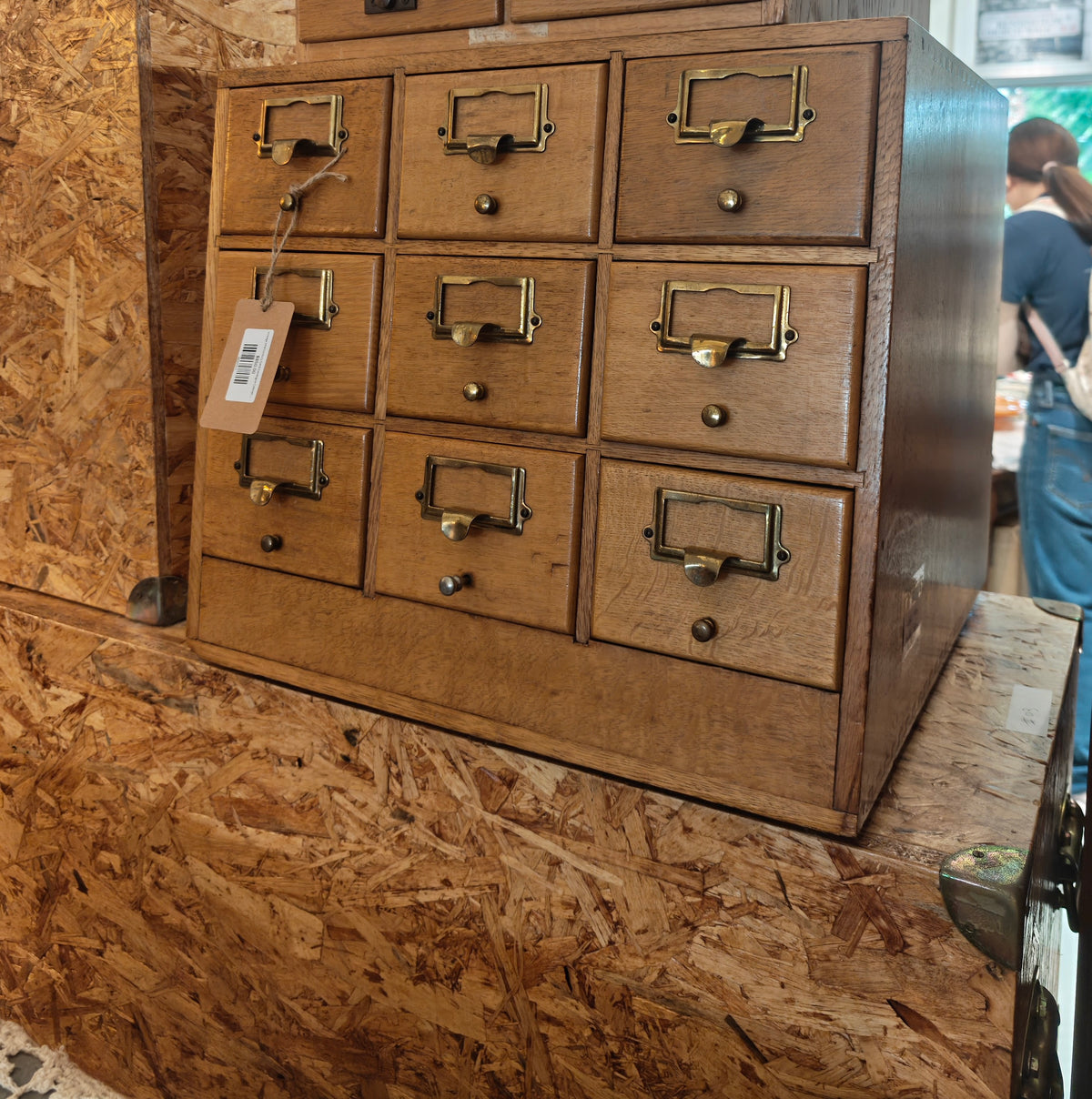 A vintage 9-drawer wooden card file cabinet with brass handles, shown in a workshop or antique store setting with OSB board walls.