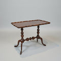 A dark brown carved wooden end table with a scalloped apron-front top and turned legs, positioned in a showroom between red leather armchairs.