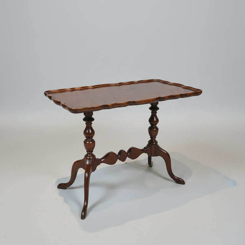 A dark brown carved wooden end table with a scalloped apron-front top and turned legs, positioned in a showroom between red leather armchairs.