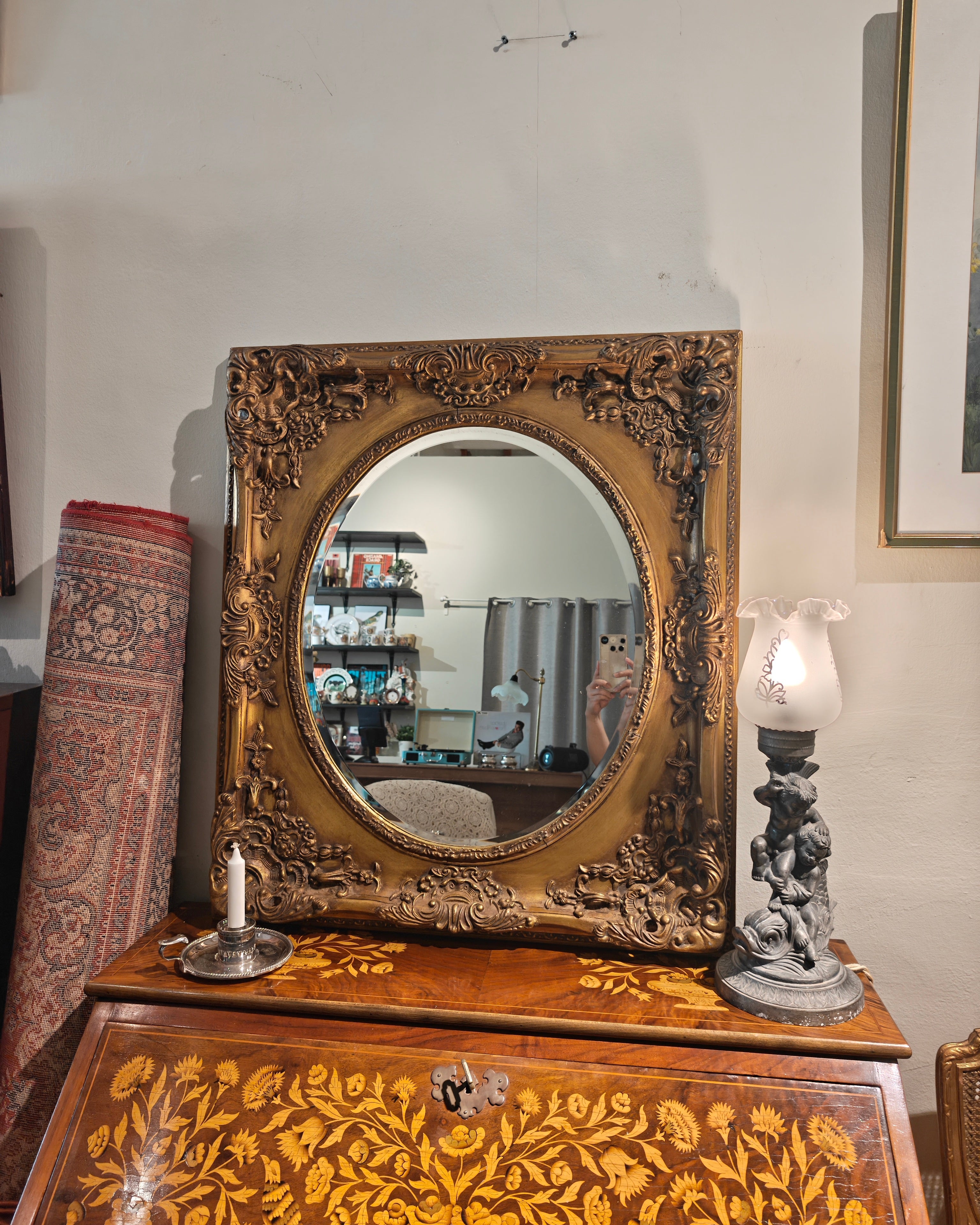 An ornate oval mirror with a carved gold frame sits on top of a wooden writing desk decorated with floral inlay. The scene is photographed in a home interior, with a wall, hanging picture frame, rolled rug, candle holder, and lamp visible.