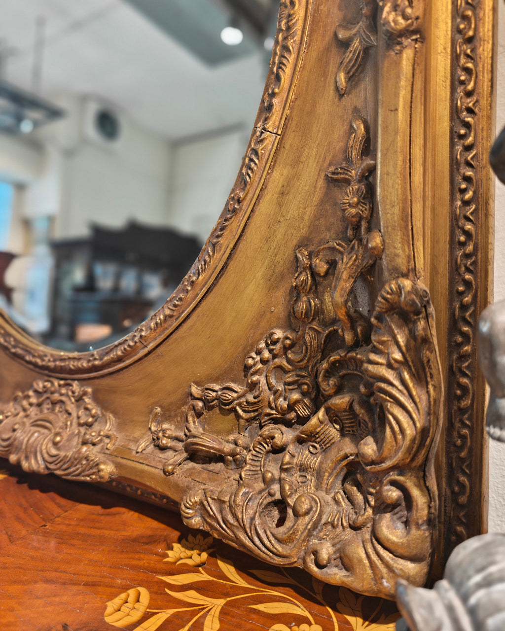 Close-up of an ornate carved wooden mirror frame in a warm golden-brown finish, resting on a patterned wooden surface with a blurred indoor room in the background.