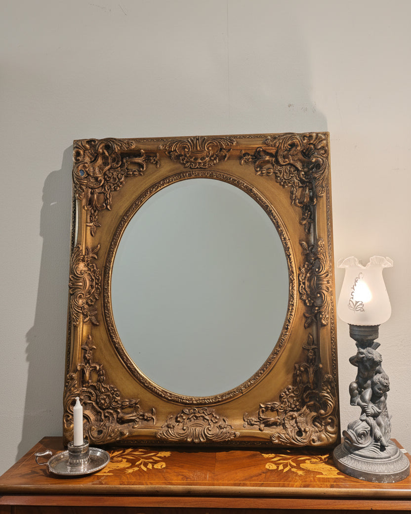 An ornate oval mirror with a heavily carved, baroque-style gold frame, displayed on a wooden console table next to a lit table lamp and a patterned textile.