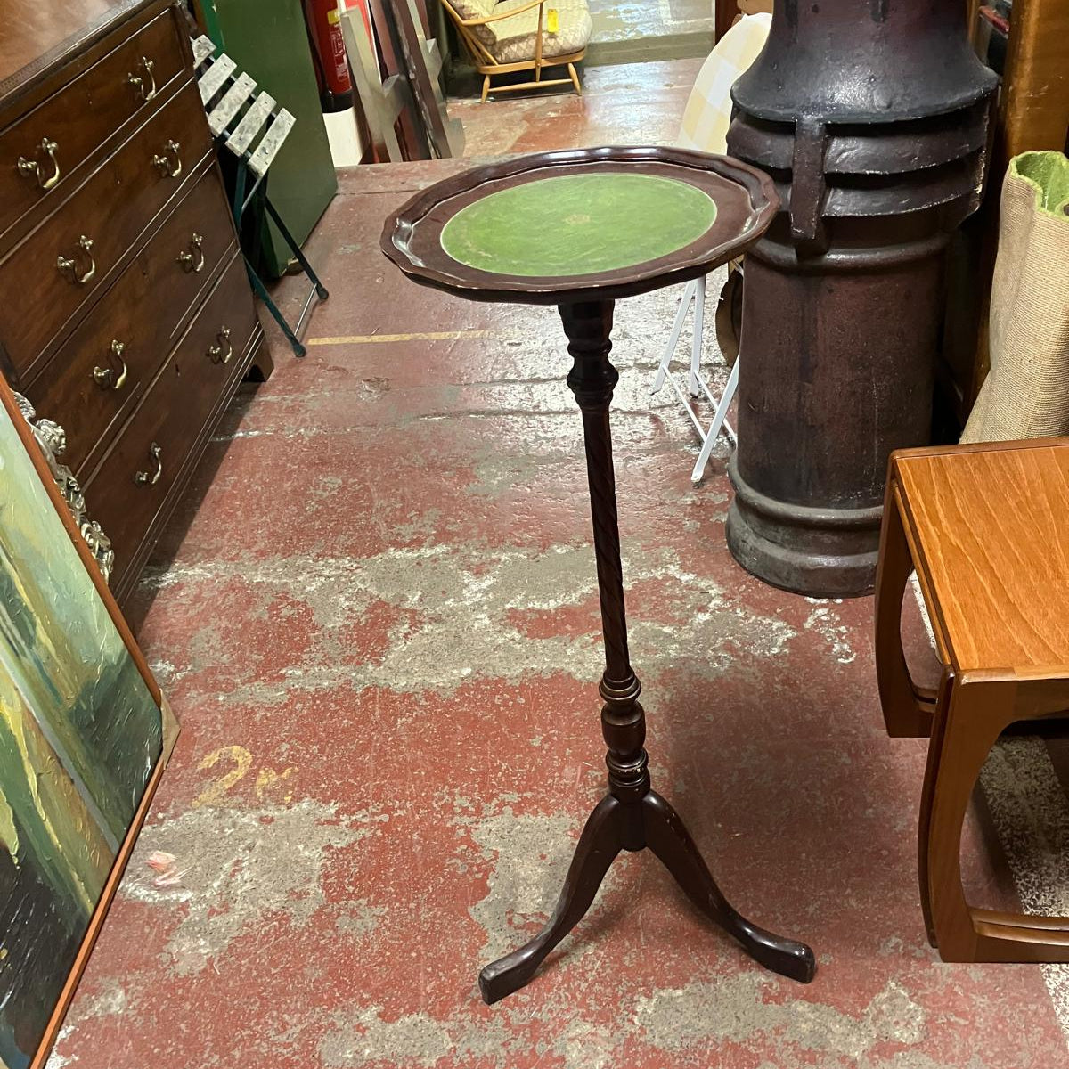 A vintage dark wood pedestal table with a round green upholstered top, standing on a three-legged base in what appears to be a furniture or antique store.